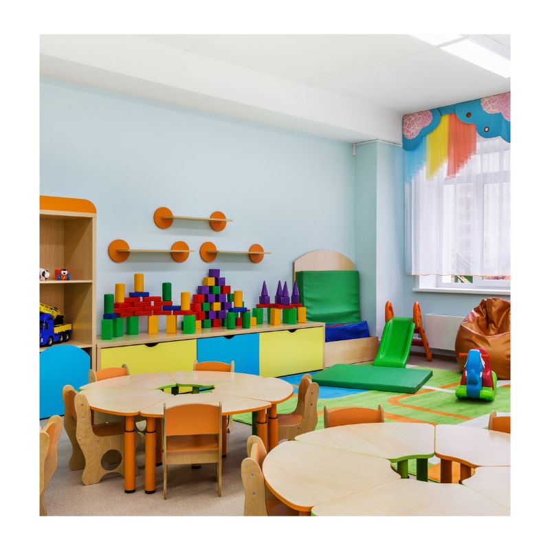 An empty medium-sized preschool classroom featuring round tables, small chairs, and an early childhood learning corner with a beanbag, slide, and educational manipulatives."