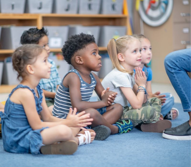 A group of preschoolers sitting together on a classroom floor listening closely as their early childhood teacher reads aloud from a book, demonstrating interactive print tracking and foundational listening comprehension skills during a structured group lesson.