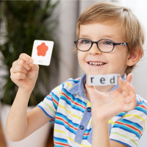 A smiling child sitting at a desk participating in a multi-sensory, play-based learning activity, holding a picture card of a red star in one hand and matching it to dice that read the word 'red' in the other hand to build interactive early literacy skills.