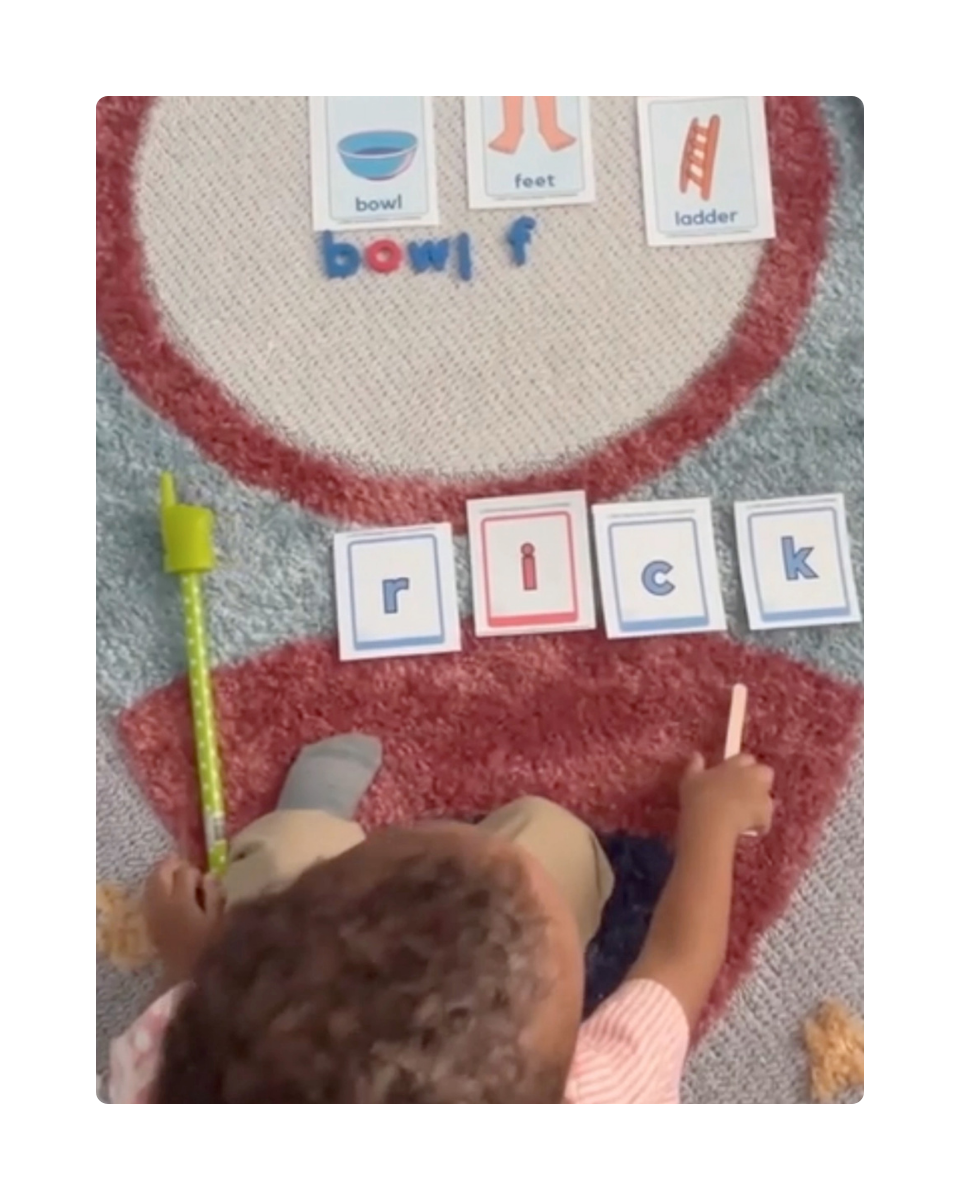 A close-up shot of a young student using a pointer stick to blend three- and four-letter CVC words using red and blue phonics blending cards. The child is practicing early reading skills on a colorful spaceship rug surrounded by magnetic alphabet letters, and additional learning cards.