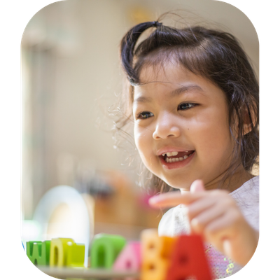 A smiling young Chinese girl sitting at a table actively pointing to alphabet letters, illustrating a screen-free, play-based early childhood phonics lesson designed to build kindergarten readiness.