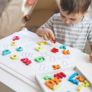 A young boy sitting at a table matching colored magnetic letters to large block letters written on a white sheet, illustrating a hands-on phonics first foundation lesson designed to teach a child to read through print recognition.