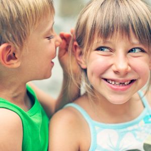 A young boy whispering into a little girl's ear as she listens closely, illustrating the early literacy concept that reading starts with the ears through phonological awareness and auditory sound recognition