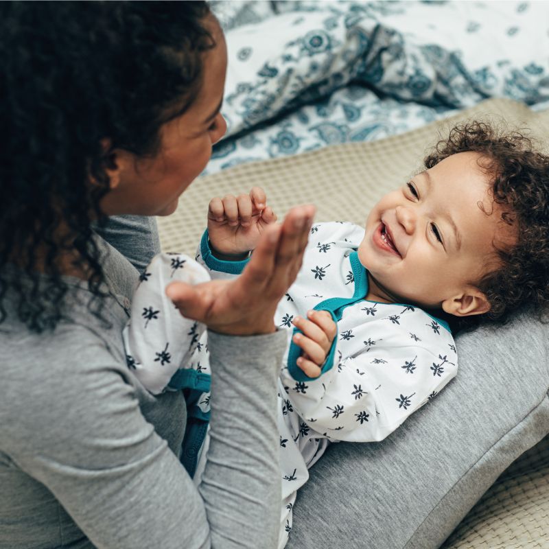 A mother sitting on the floor holding her smiling 12-month-old child on her lap while playing and singing, demonstrating active early brain development and responsive parenting methods that wire crucial neural sound connections.