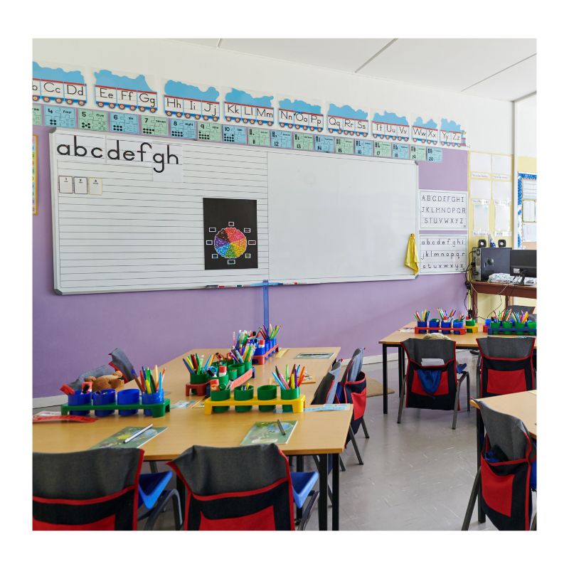 n elementary school classroom set up for early literacy instruction, featuring individual student desks grouped into centers, an alphabet train banner on the wall, and a whiteboard with handwriting lines displaying lowercase letters a through f.