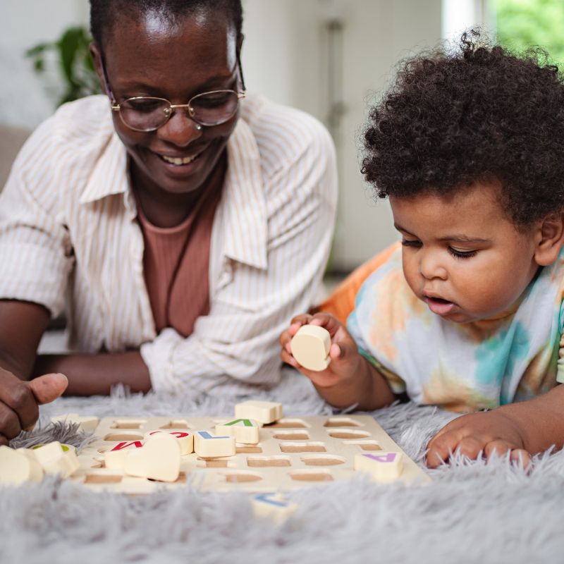 A mother and toddler sitting on the floor together working on an alphabet puzzle, showcasing a 3-minute micro-lesson from the kindergarten curriculum where the child finds and matches letters to build early print recognition.